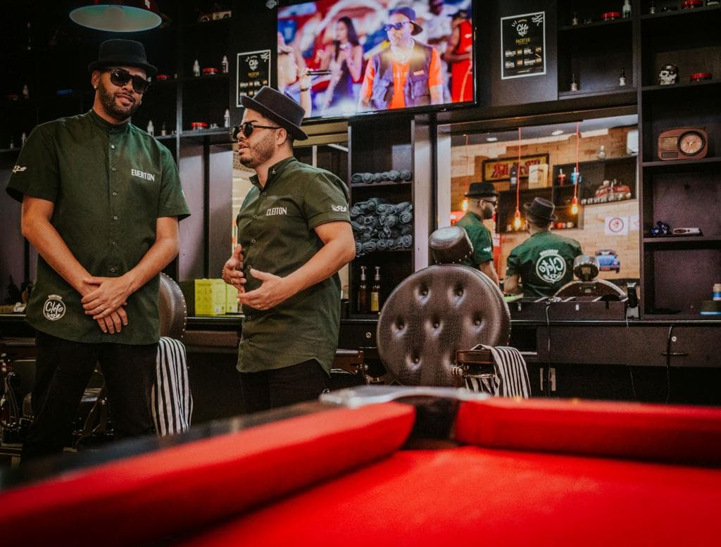 Dois barbeiros de farda verde da Cleto conversando no salão, com a mesa de sinuca vermelha em primeiro plano